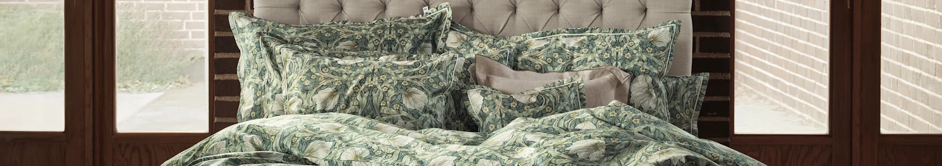 A bed with a tufted headboard and green and cream floral patterned duvet and pillows, beside windows with wooden frames.