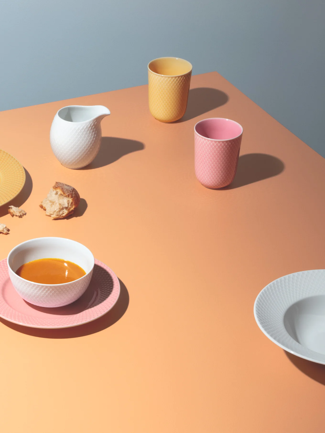 Table setting with white soup bowl on pink plate, white pitcher, yellow and pink textured cups, and bread on an orange surface.