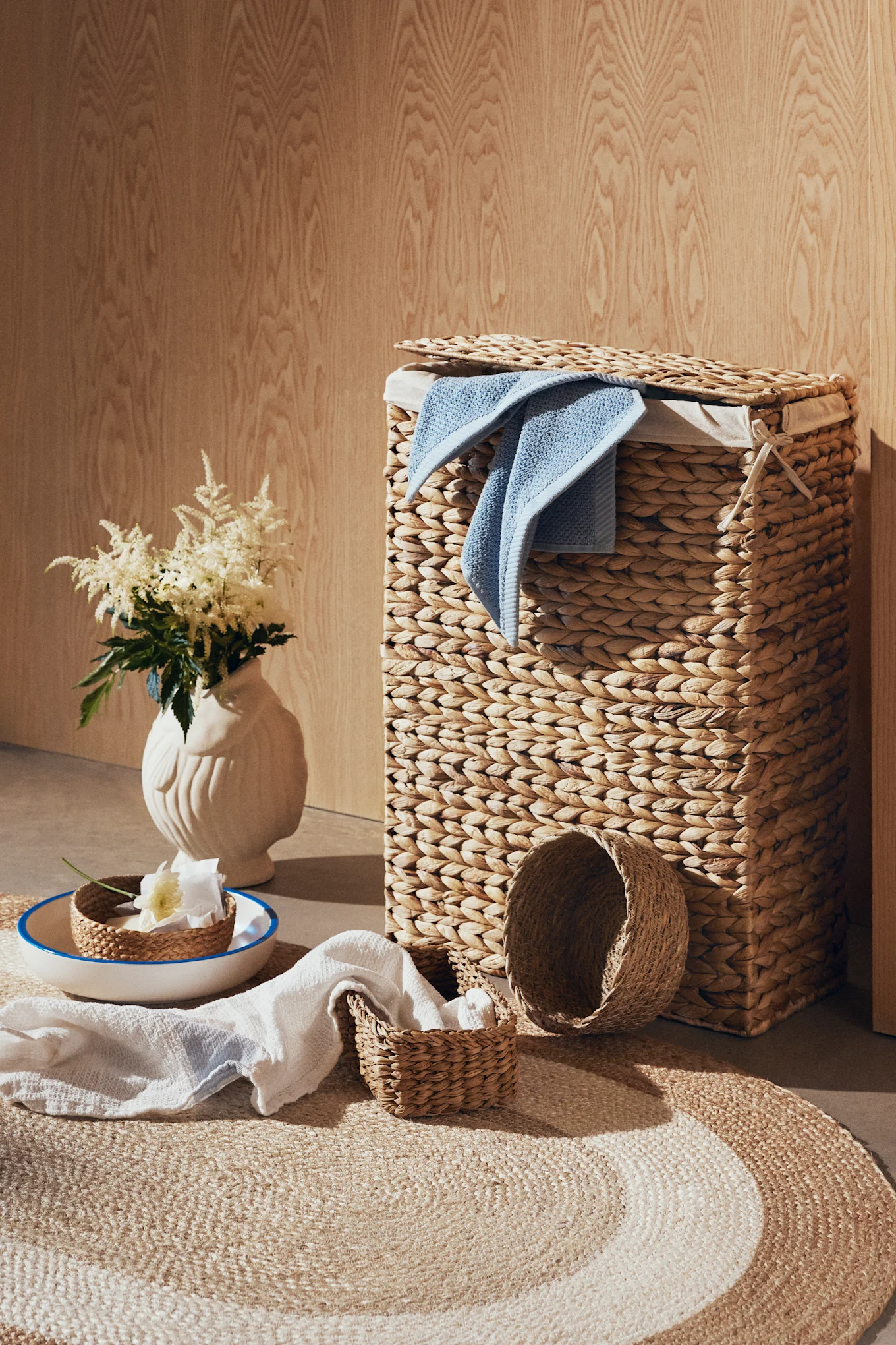 Woven laundry hamper with blue towel, small baskets, white flowers, and a round rug.