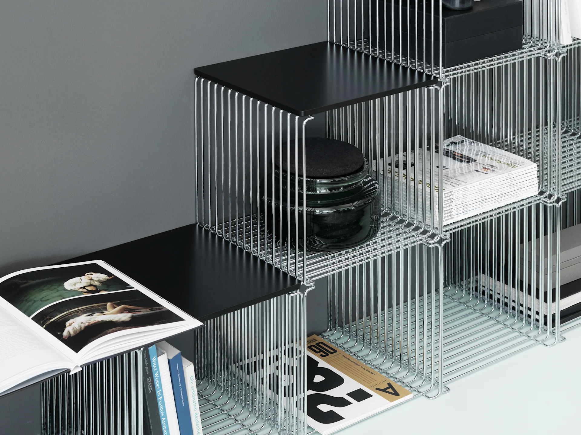 Modular chrome wire shelving unit with black shelves, holding books, magazines, a black box, and a dark jar against a grey wall.