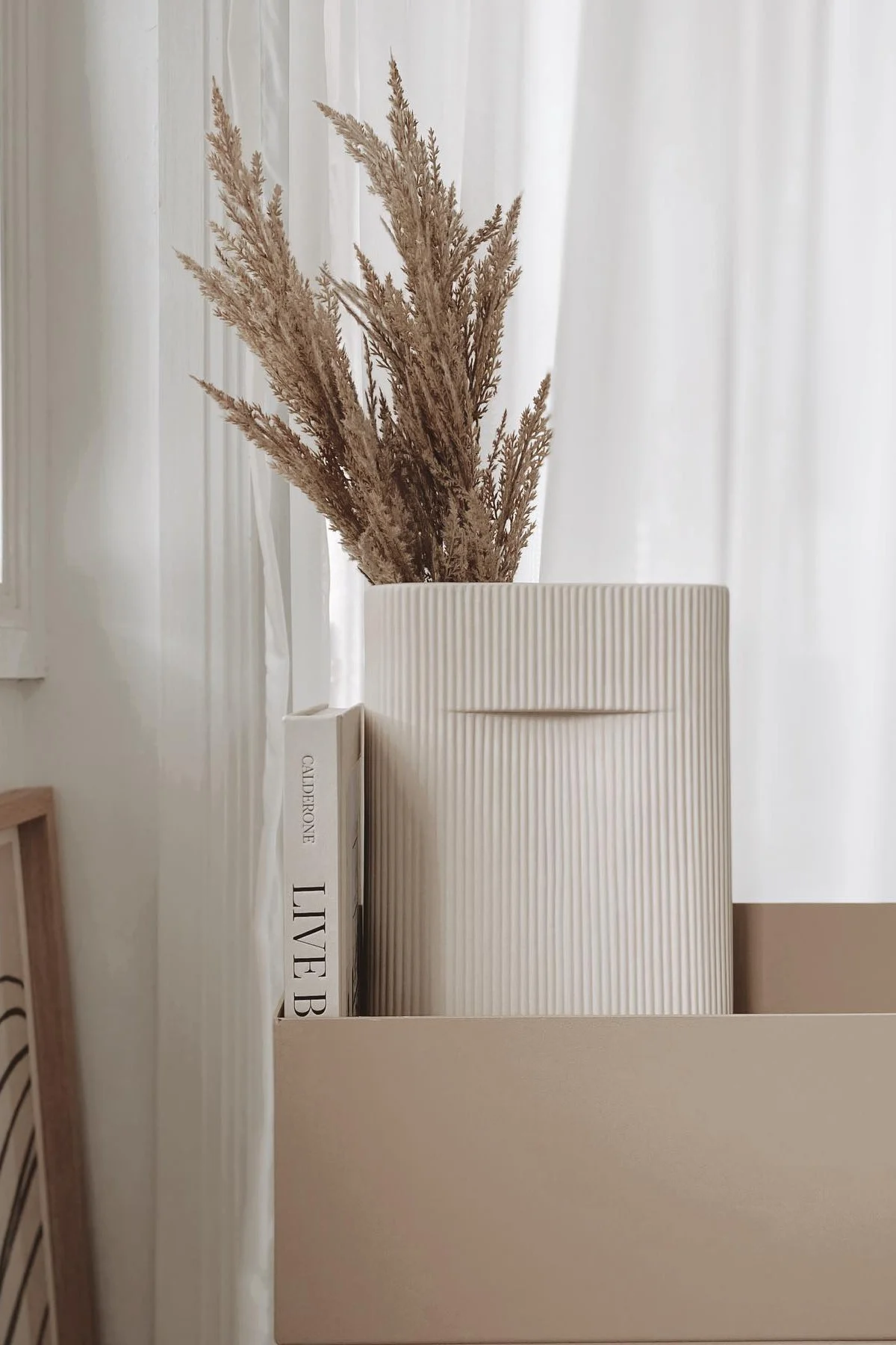 Dried pampas grass in a ribbed beige vase next to a book on a beige shelf.