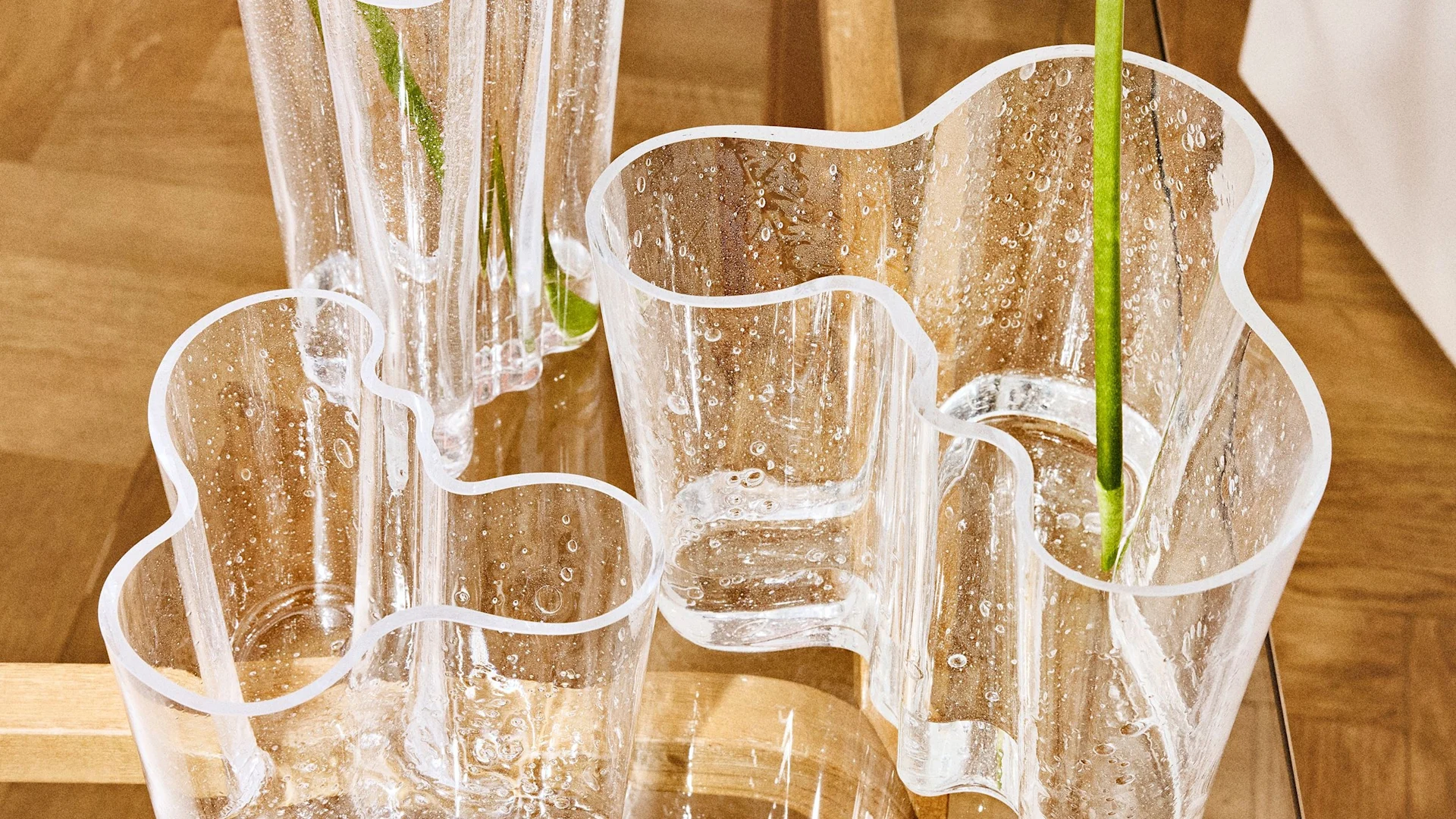 Several clear glass vases with organic wavy shapes and embedded air bubbles, holding green stems, on a wooden surface.