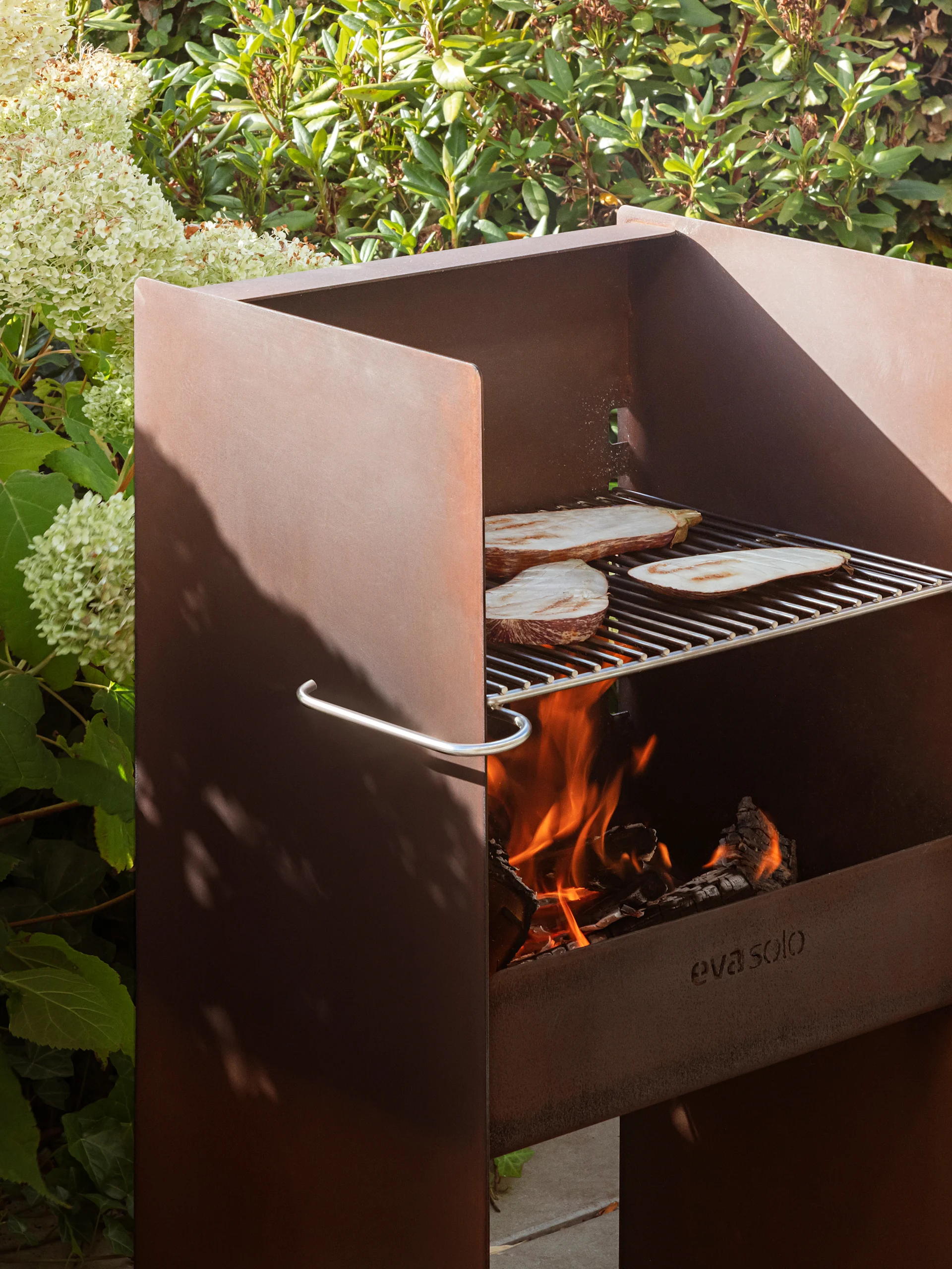 Rusted metal outdoor grill by Eva Solo with eggplant on the grate, fire burning, and lush foliage.