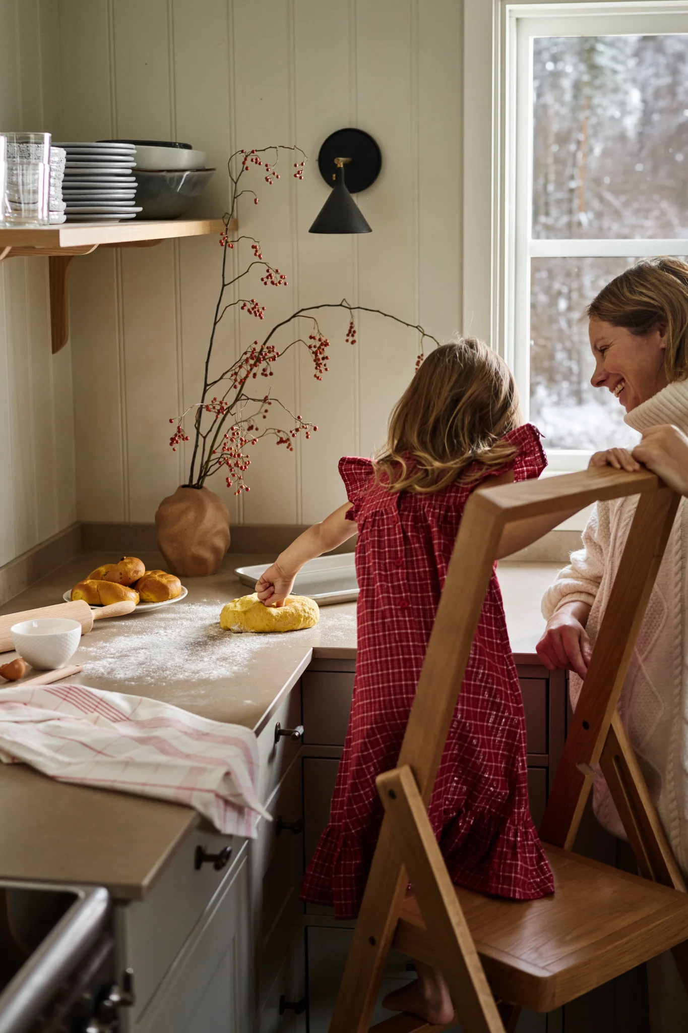 Child in a red dress on a stool kneading yellow dough with an adult smiling next to them in a flour-dusted kitchen.