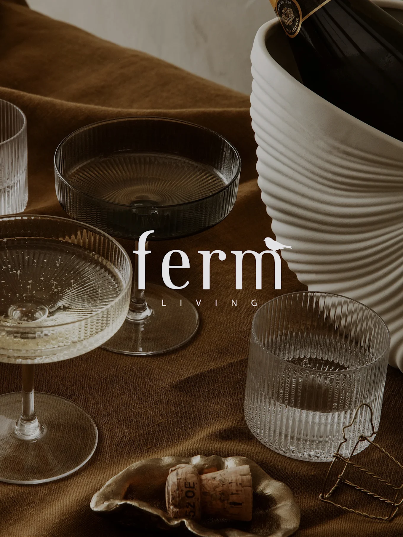 Champagne bottle in ribbed cooler, champagne coupe, ribbed tumbler, and cork on a dish, on brown fabric.