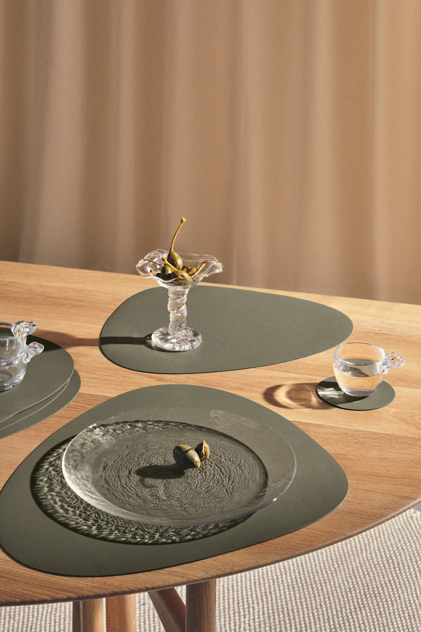 A modern table setting with green placemats, clear glass plates, a bowl of caper berries, and a glass of water on a light wooden table.
