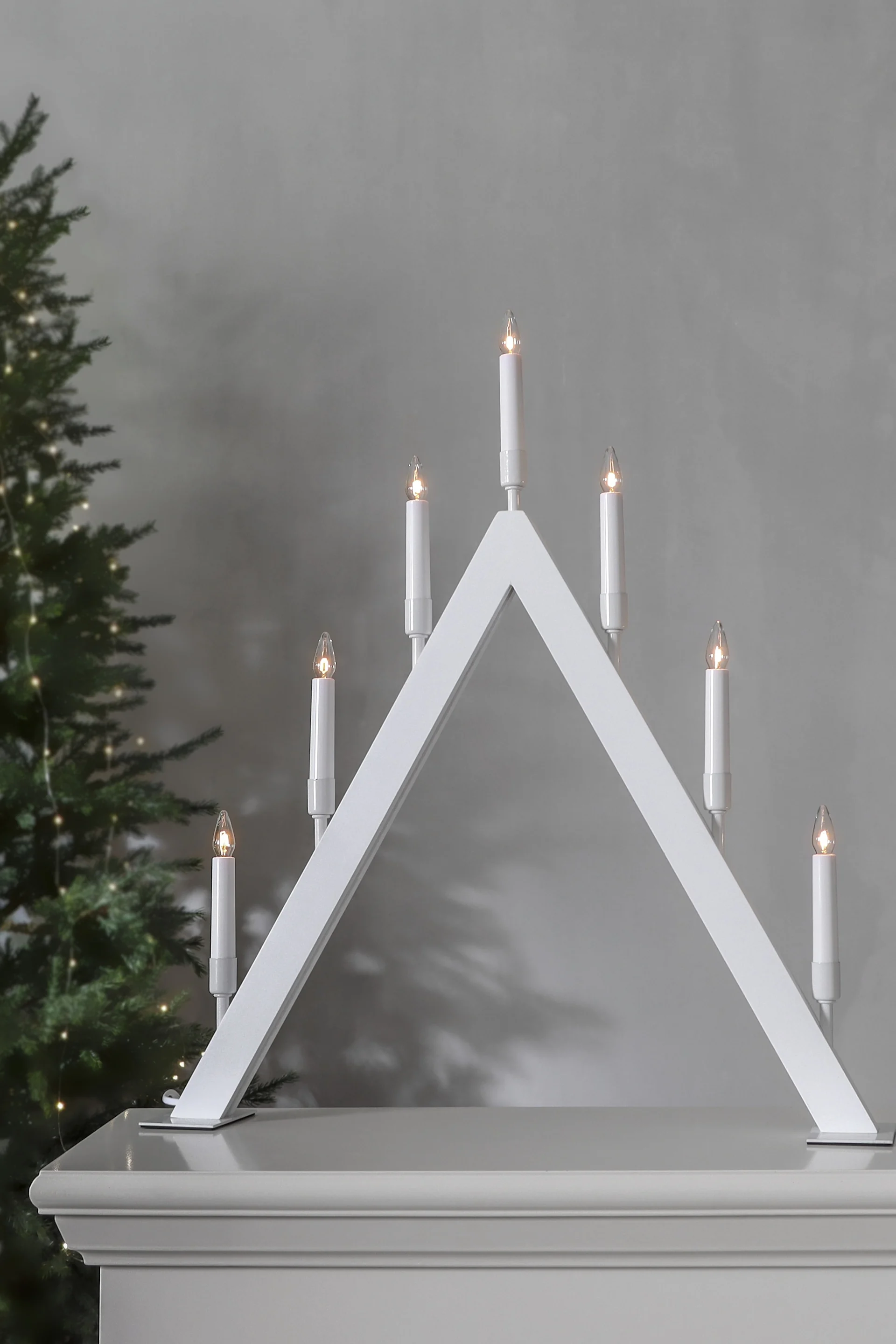 A white, triangular candle holder with seven lit electric candles sits on a white surface, with a blurred Christmas tree in the background.