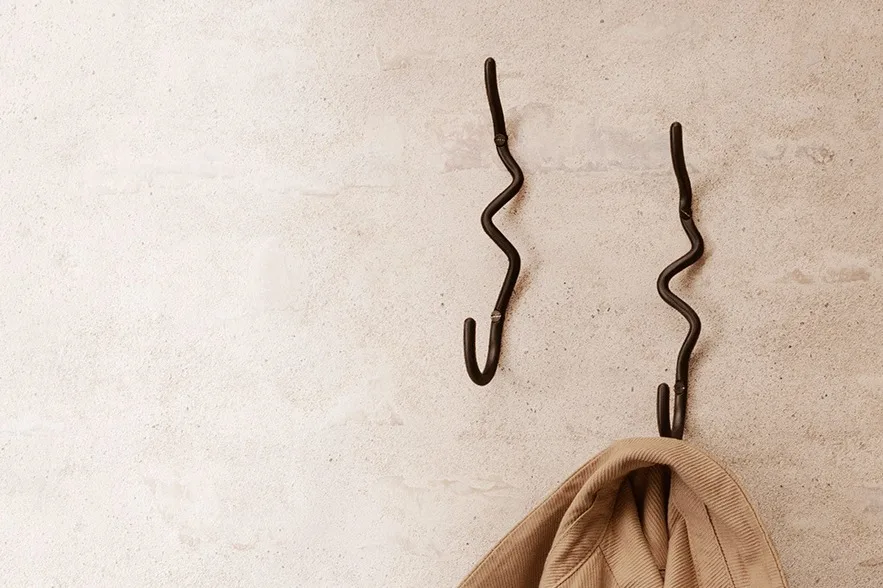 Two wavy dark metal wall hooks on a light textured wall, with a beige jacket hanging on one.
