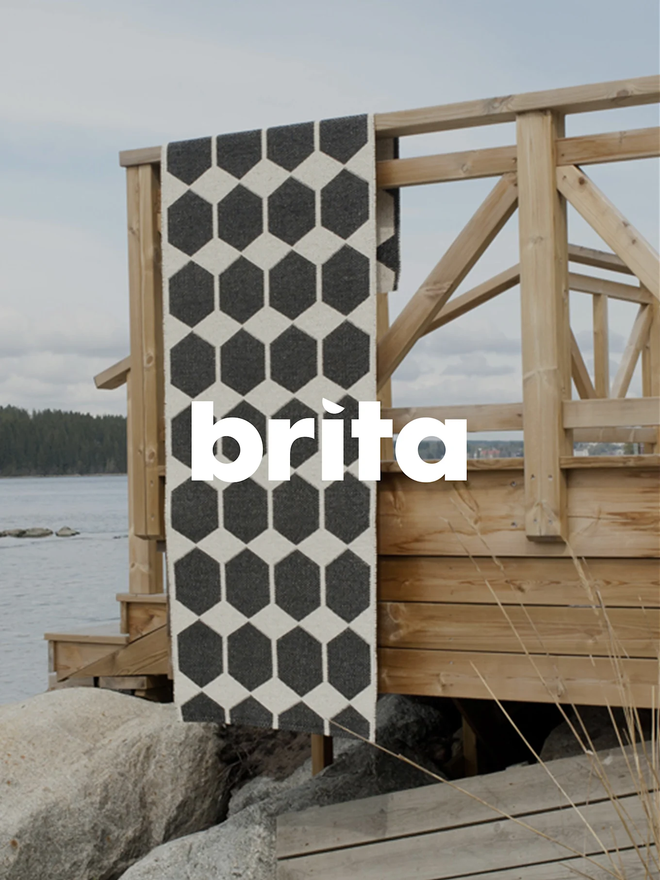 A black and off-white hexagonal patterned runner rug draped over a wooden railing by a calm body of water with rocks.