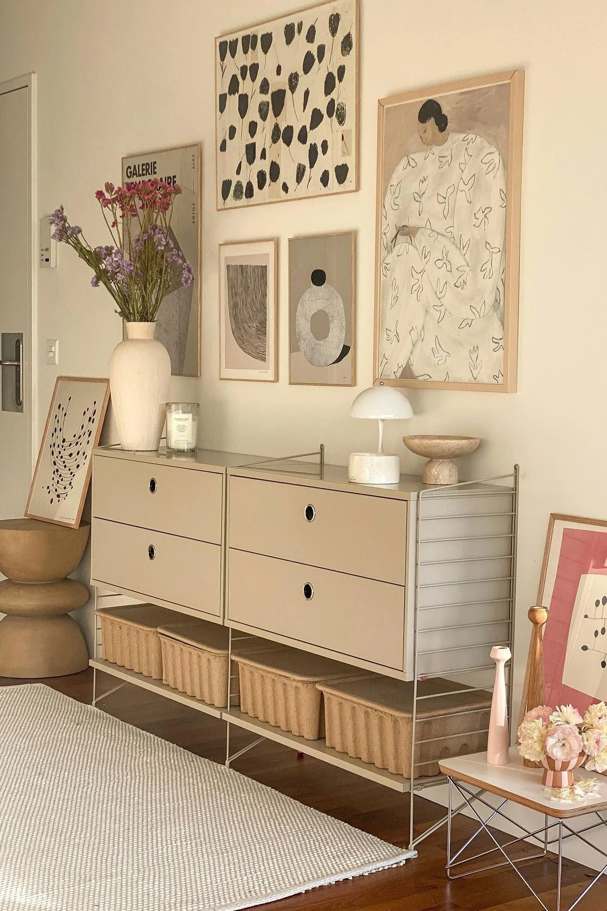 A modern room with a beige modular cabinet, drawers, and storage baskets. A vase of flowers, lamp, and abstract art adorn the space.