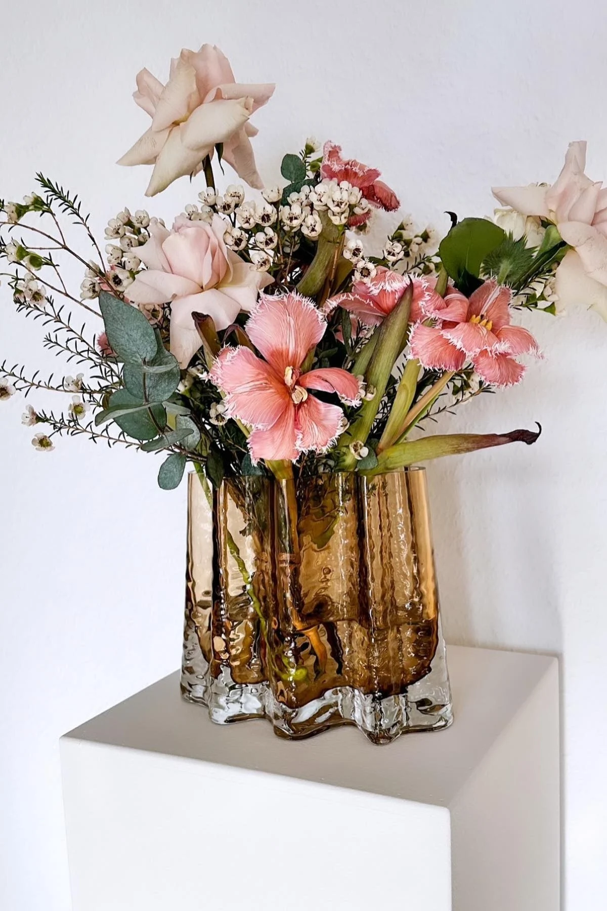 A bouquet of pink roses, fringed tulips, and small white flowers in a textured bronze vase on a white pedestal.