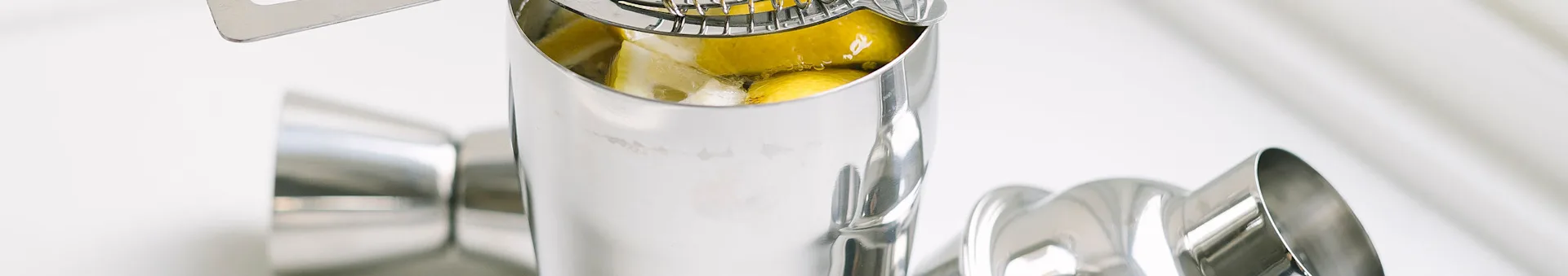 Close-up of a stainless steel cocktail shaker with lemon wedges and liquid inside, surrounded by other bar tools.