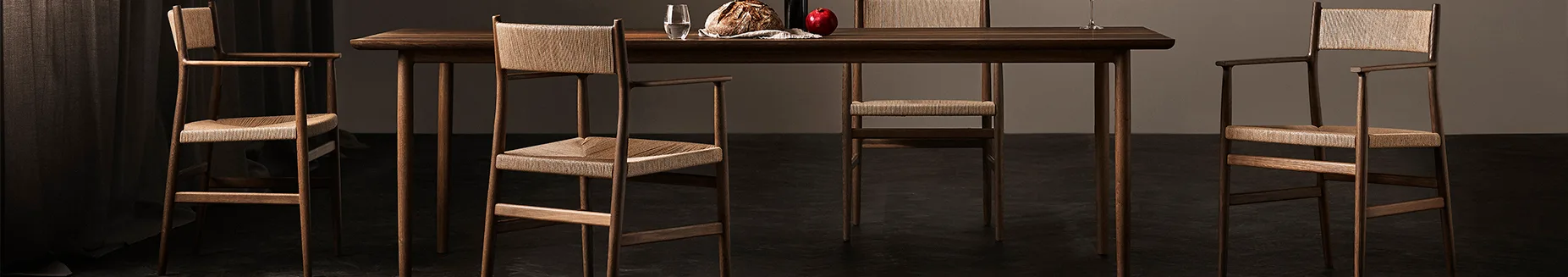 Dark wooden dining table with four chairs featuring woven seats and backs, set with bread, a glass of water, and an apple.