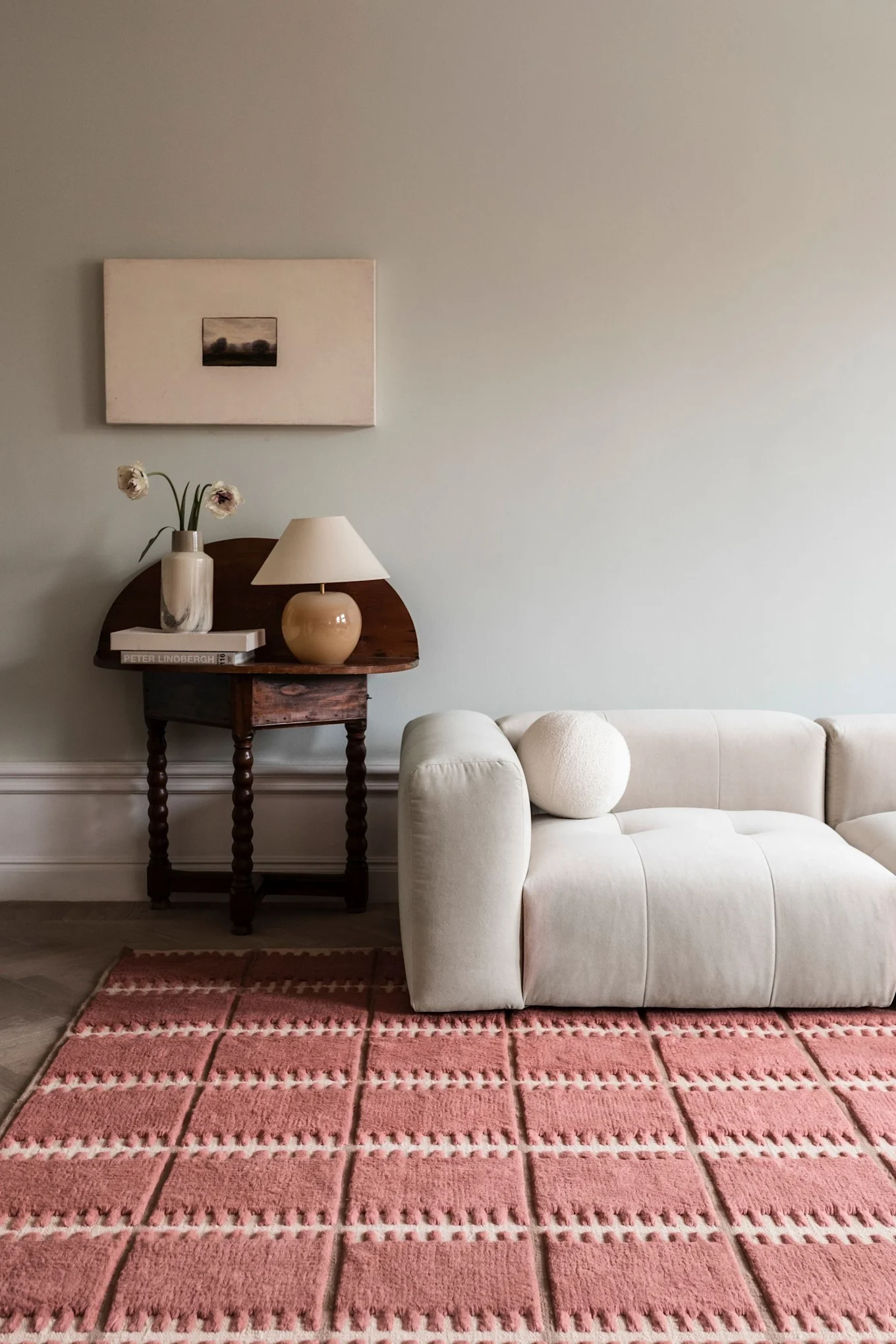 A cream modular sofa with a round cushion, a wooden table with a lamp, vase, and books, and a textured pink and white rug.