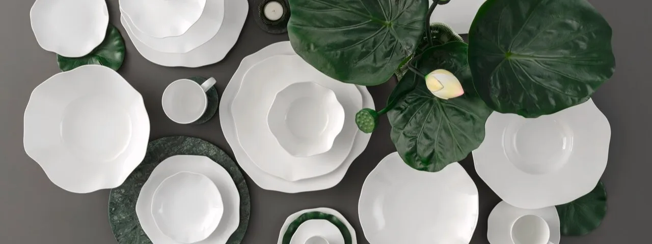 An overhead flat lay of white ceramic dinnerware with wavy edges, arranged with green leaves and marble coasters on a grey background.