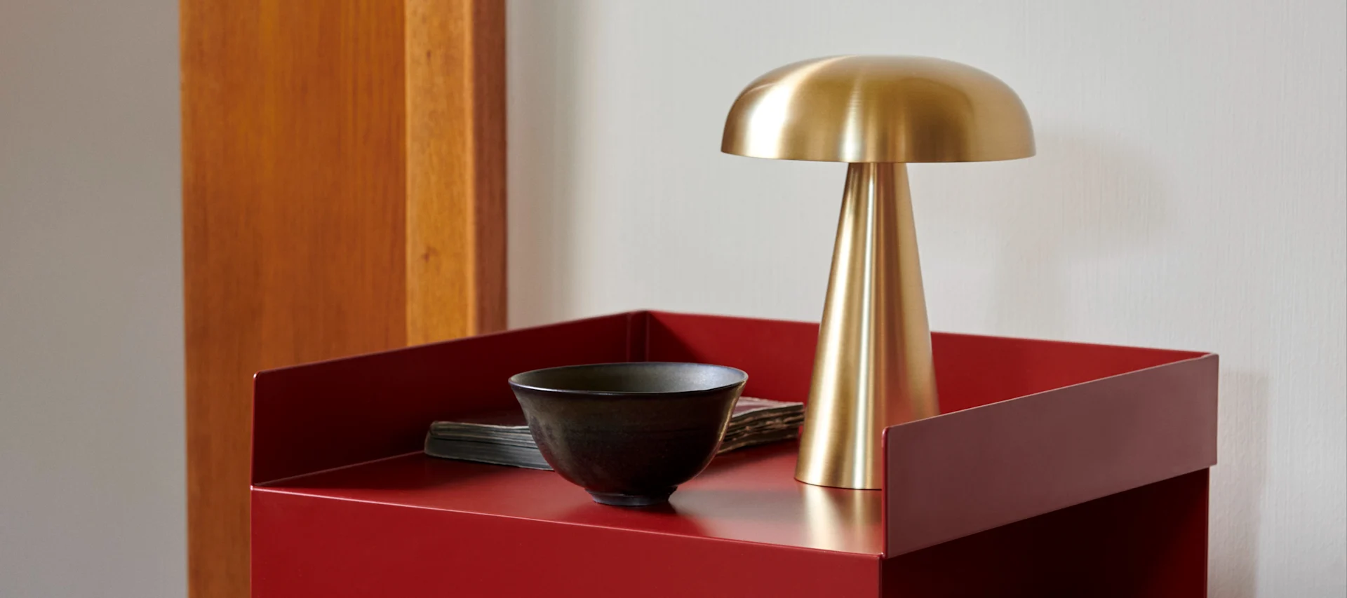 A brass mushroom lamp and a dark bowl on a red metal side table next to a wooden panel.