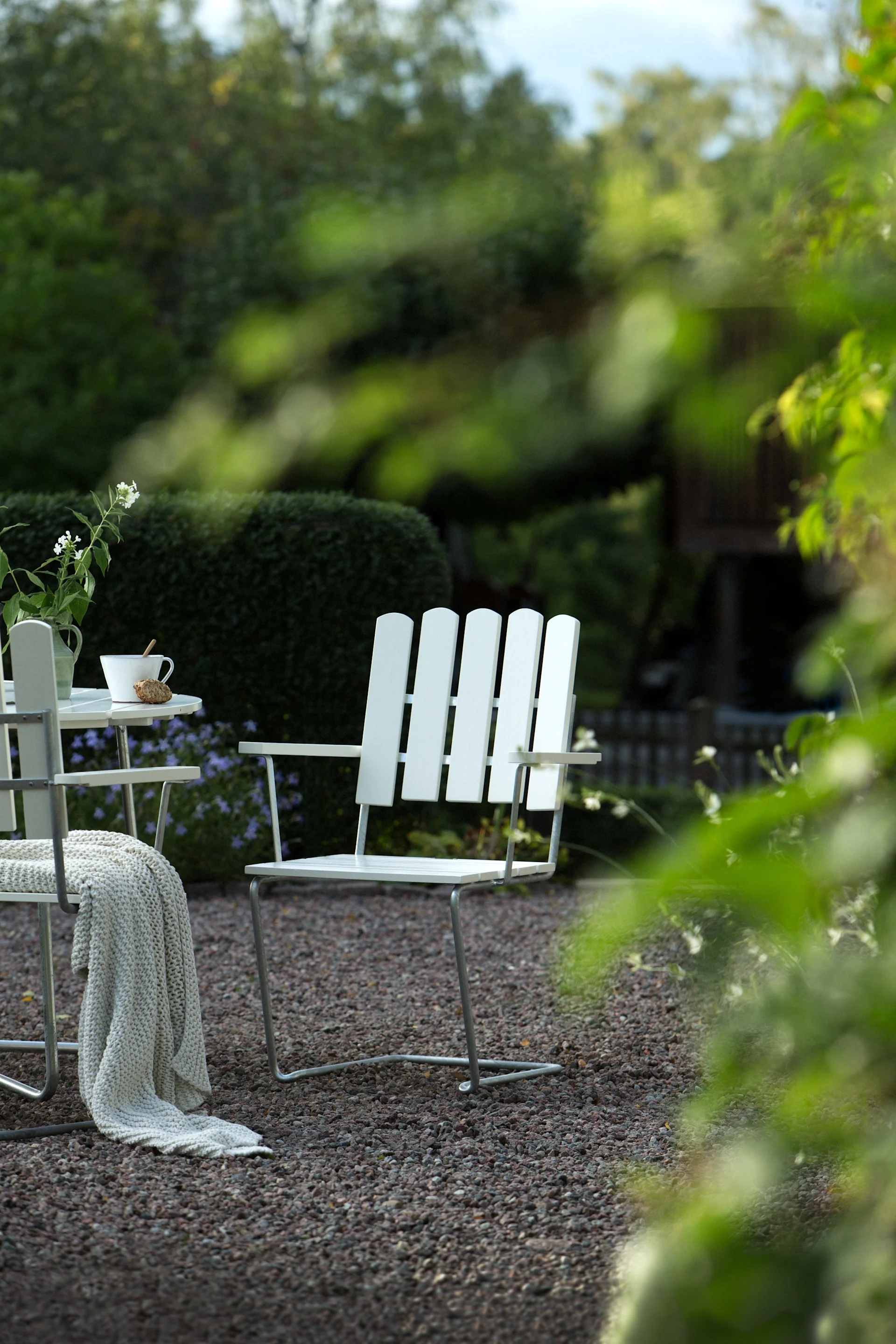 White garden chair on gravel patio. Adjacent chair has knitted blanket. Small table with flowers and cup. Lush green background.
