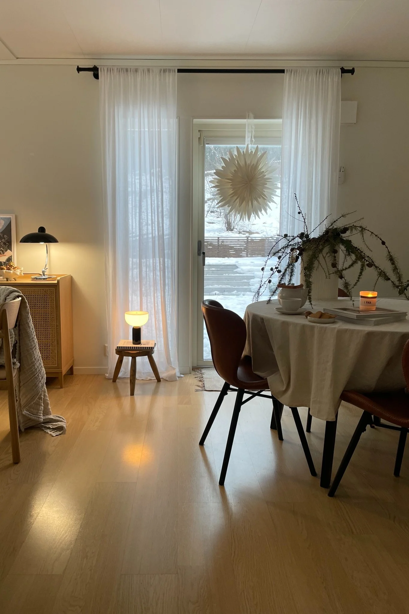 Cozy interior with a round dining table, brown chairs, and snowy window view. Table holds branches, a candle, and cookies.