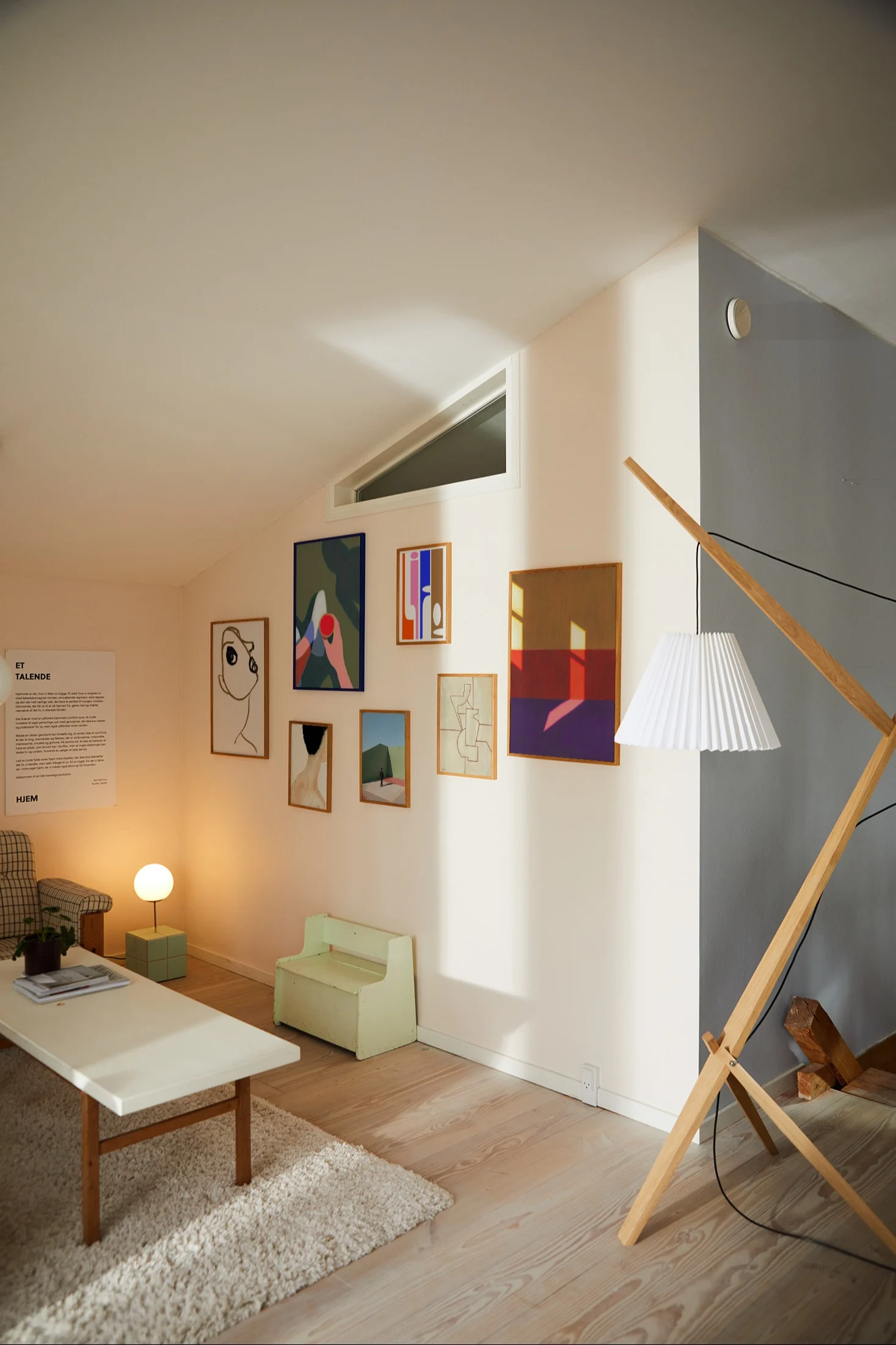 A bright living room with a gallery wall of abstract art, a white coffee table, a plaid armchair, and a wooden floor lamp.