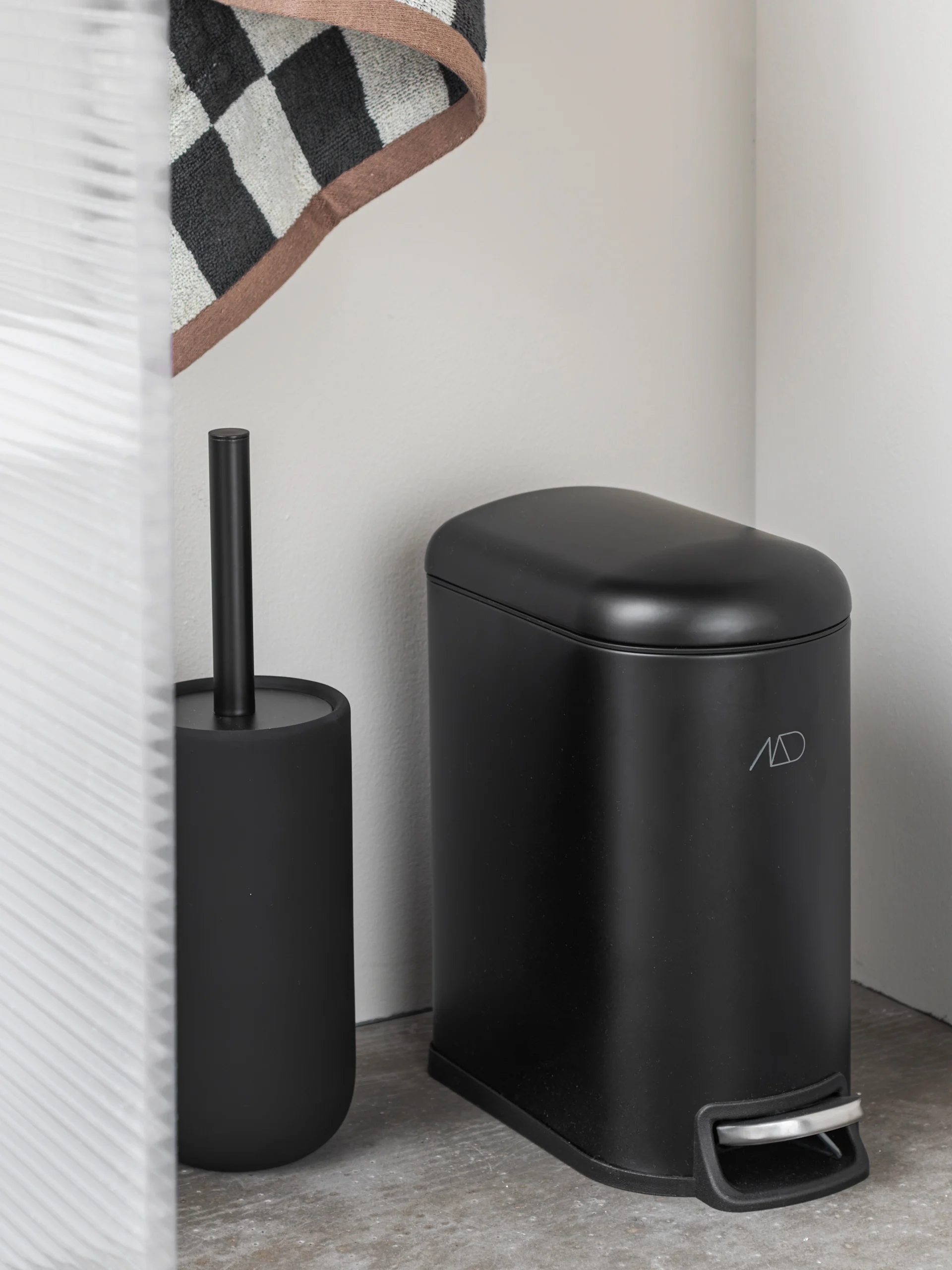 A matte black toilet brush and holder next to a matching black pedal bin with a silver foot pedal, against a white wall.