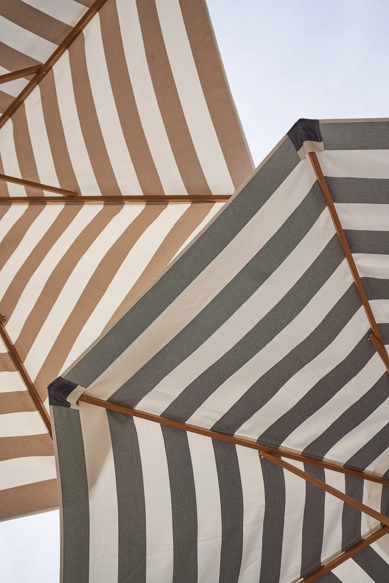 View from below of two striped patio umbrellas against a light sky, one brown and white, the other dark grey and white.