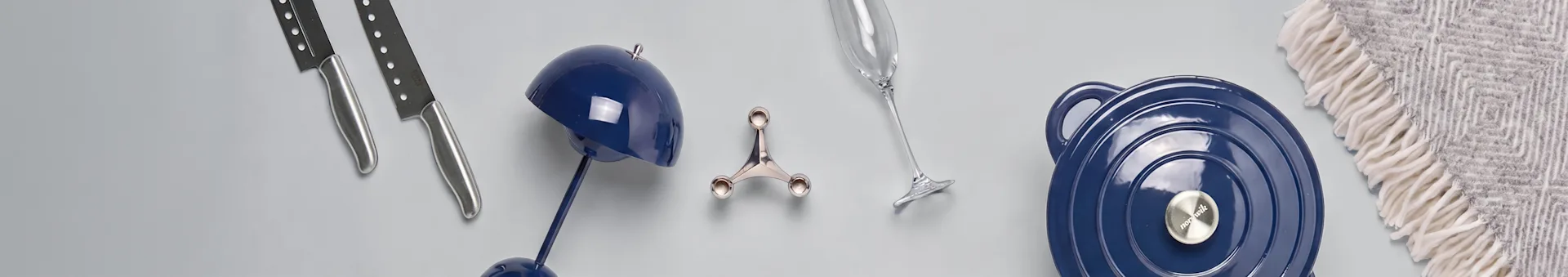 Assortment of kitchenware and home decor: two silver knives, a blue decorative lamp, a silver kitchen tool, a champagne flute, a blue lidded pot, and a patterned throw blanket.