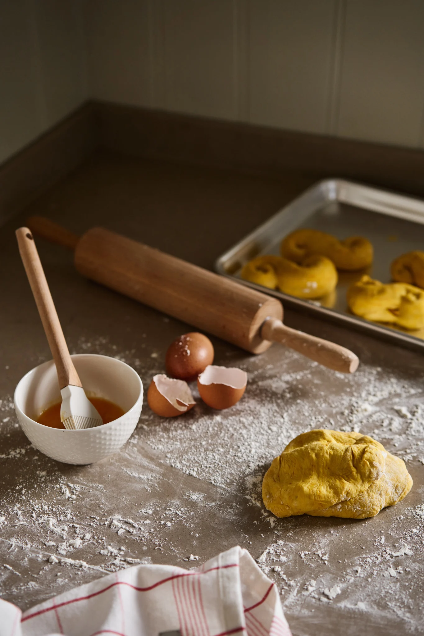 Baking scene with yellow dough, flour, rolling pin, egg wash, and saffron buns on a tray.