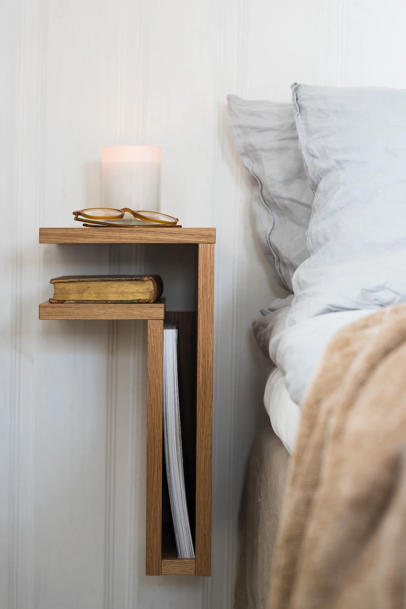 A wall-mounted wooden bedside shelf with a candle, eyeglasses, a book, and a magazine next to a bed with blue bedding.