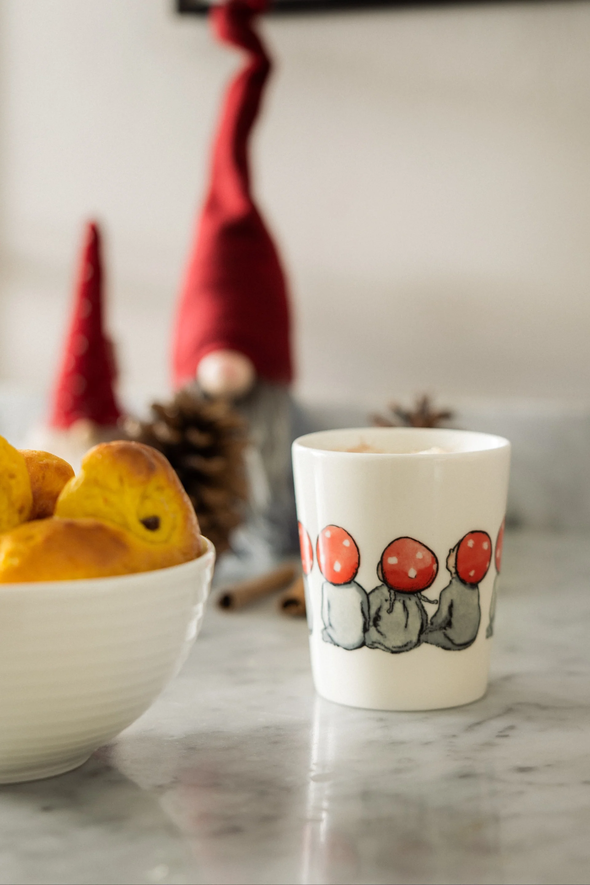 Saffron buns in a white bowl, next to a mug with mushroom gnomes, and blurred holiday decorations.