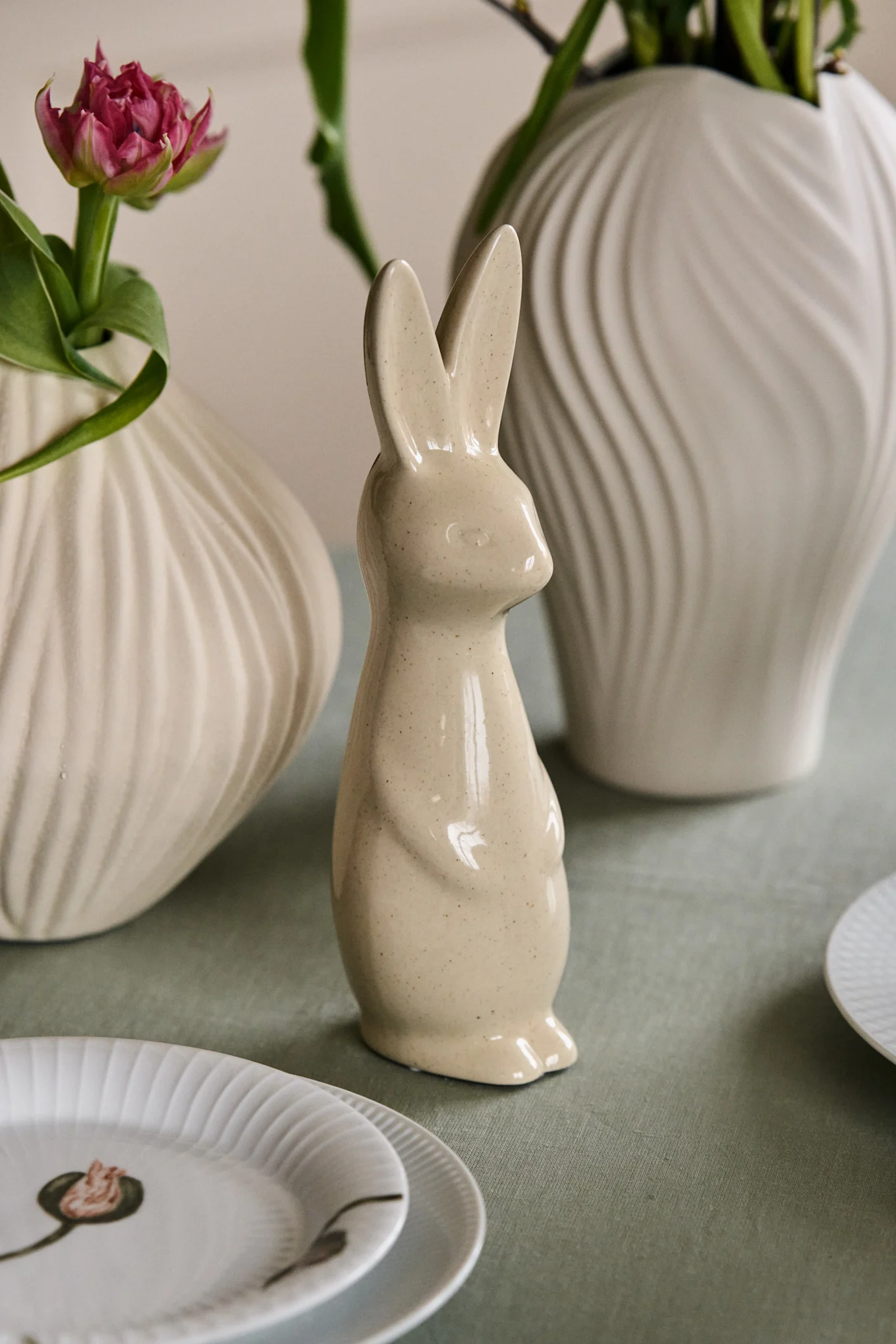 A speckled ceramic bunny figurine stands on a green tablecloth, flanked by ribbed vases with a pink tulip and green leaves, with white plates in the foreground.
