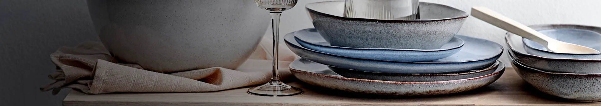 Stacked rustic ceramic bowls and plates in blue and grey tones on a wooden surface, with a glass and napkin.