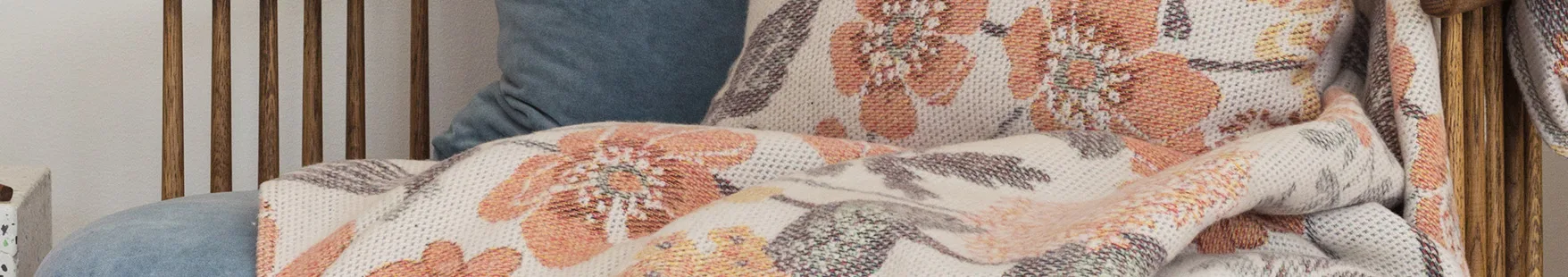 Close-up of a cream-colored throw blanket and pillow with an orange and green floral pattern on a chair.