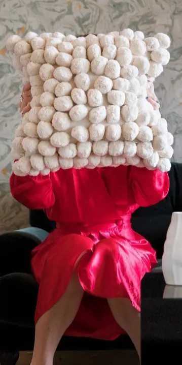 Person in a red satin dress holding a textured cream pillow with pom-poms.