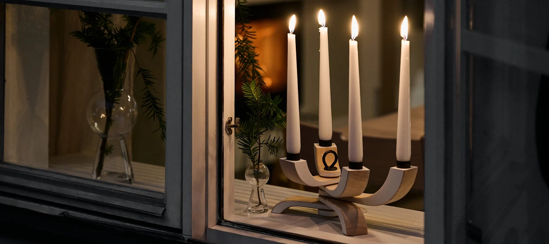 Four lit candles in a modern wooden candelabra on a window sill at night, with green branches in a vase next to it.