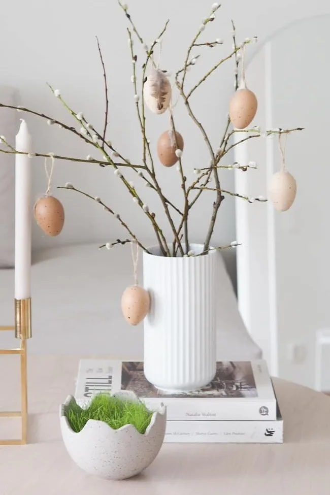 Easter decor with pussy willow branches in a white vase, hanging speckled eggs, a white candle, and a faux grass-filled eggshell bowl.