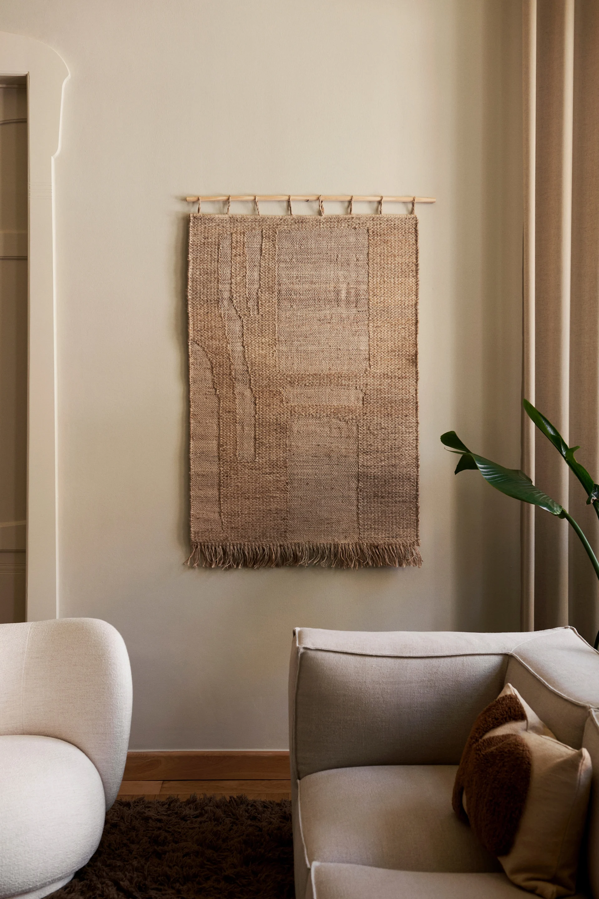 Neutral living room with a textured brown woven wall hanging, light beige sofa, off-white armchair, and green plant.