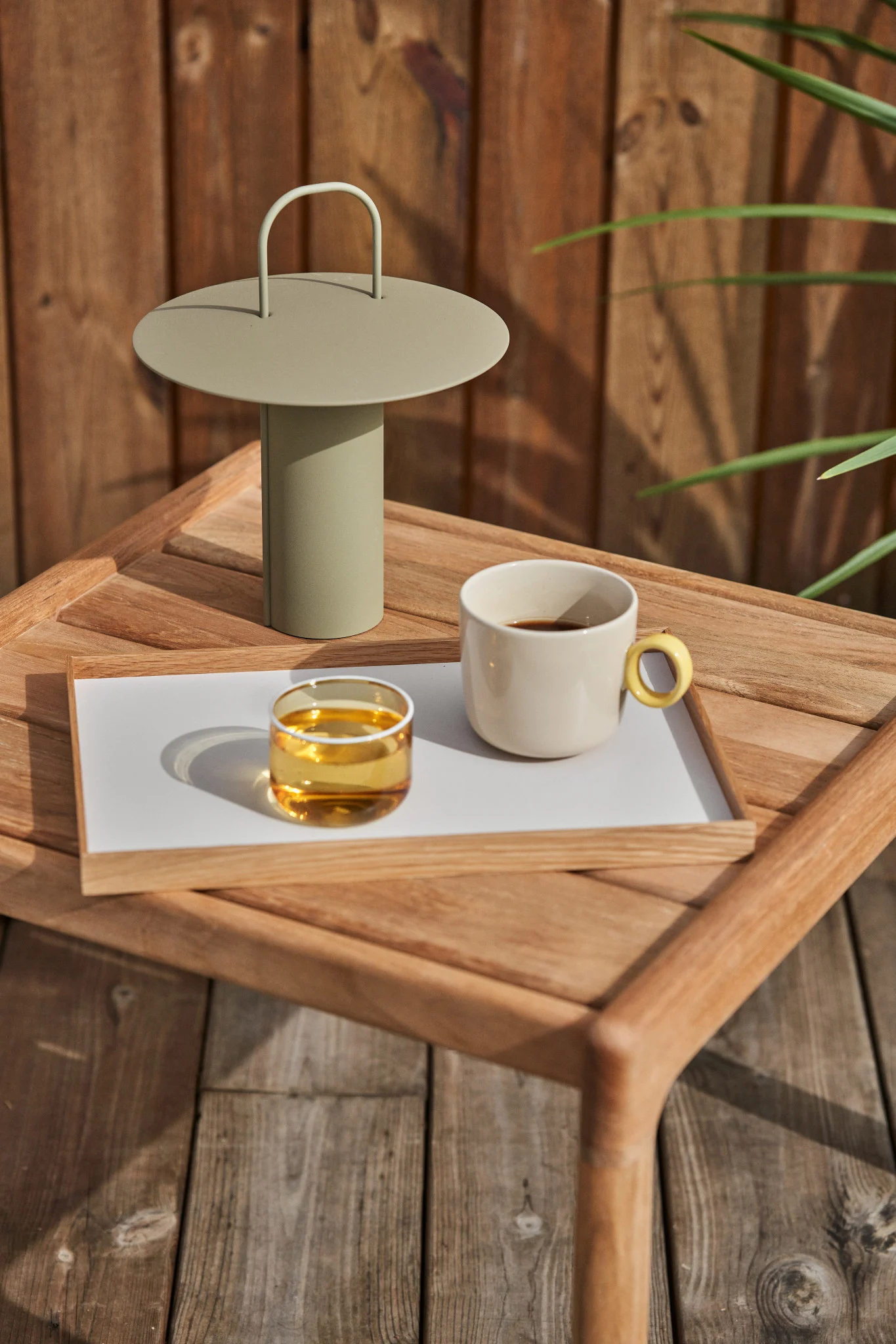 A light green portable lamp, a glass of amber liquid, and a cream mug of coffee on a wooden tray on an outdoor wooden table.
