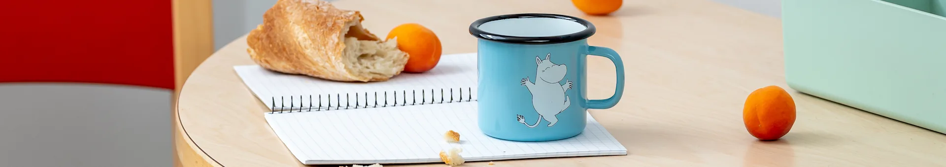 A light blue enamel mug with a Moomin character, torn bread, and two apricots on a spiral notebook on a wooden table.
