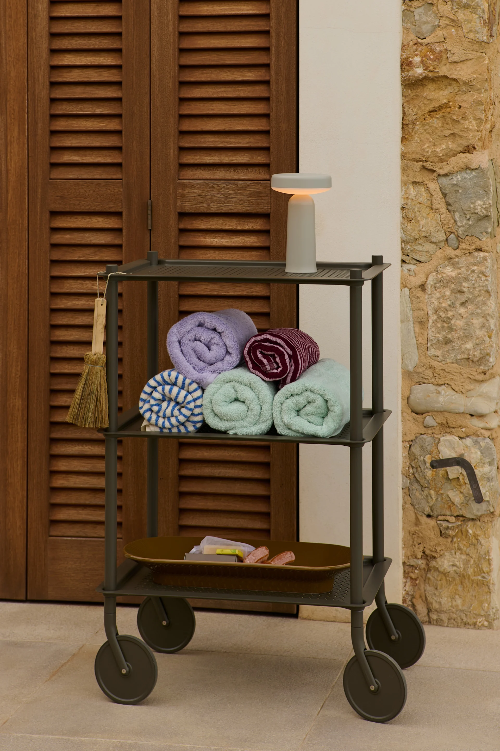 A three-tiered utility cart with rolled towels, toiletries, and a lamp next to a wooden door and stone wall.