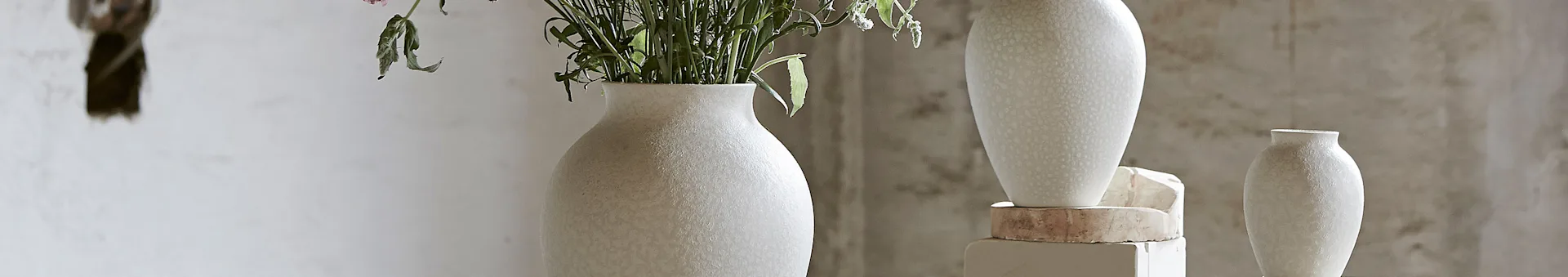 Three textured ceramic vases in light colors, two holding green plants and delicate flowers, against a textured wall.
