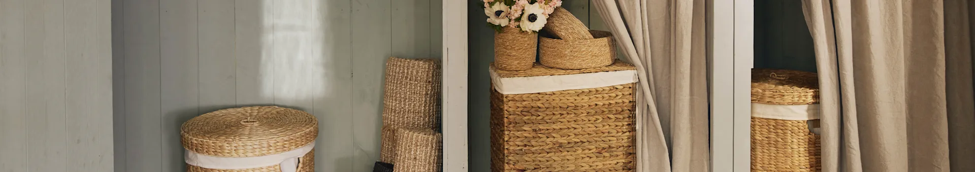 Natural woven baskets, lidded hampers, one with flowers, next to beige curtains.
