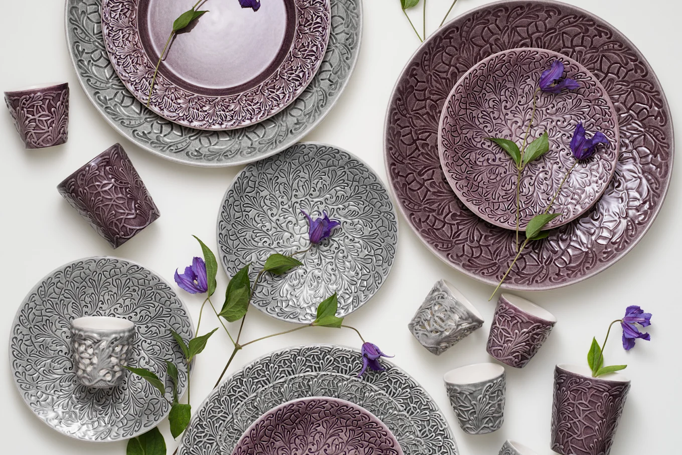 Grey and purple ceramic plates and cups with embossed floral patterns, accented by purple flowers and green leaves.
