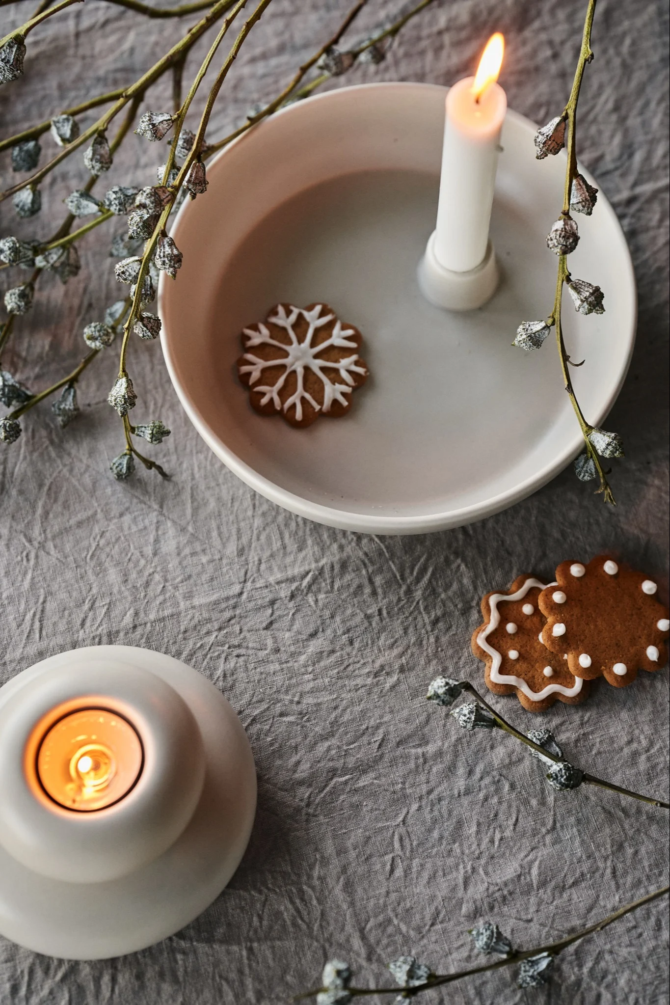 Lit candles and gingerbread cookies with white icing, surrounded by frosted branches on a grey linen cloth.