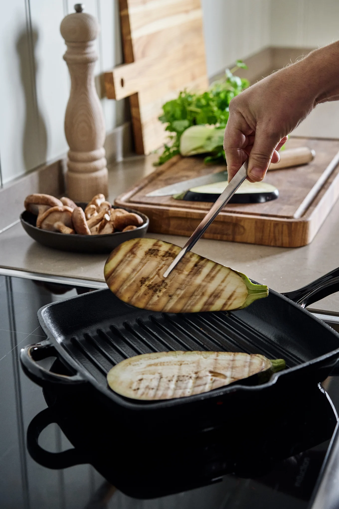 Hand flipping a grilled eggplant slice in a black grill pan on an induction hob, with mushrooms and vegetables nearby.