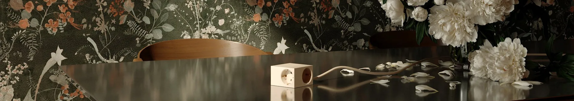 Dark floral wallpaper behind a reflective table with a cube power strip, scattered white petals, and white peonies.