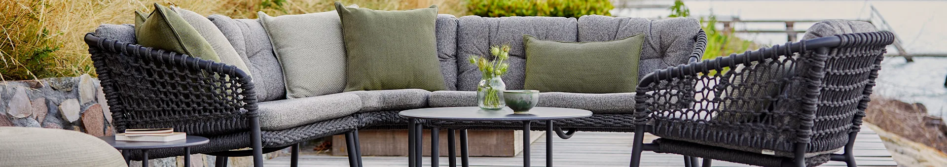 Outdoor corner sofa with dark grey woven frame, light grey cushions, and green pillows. A small table holds plants.