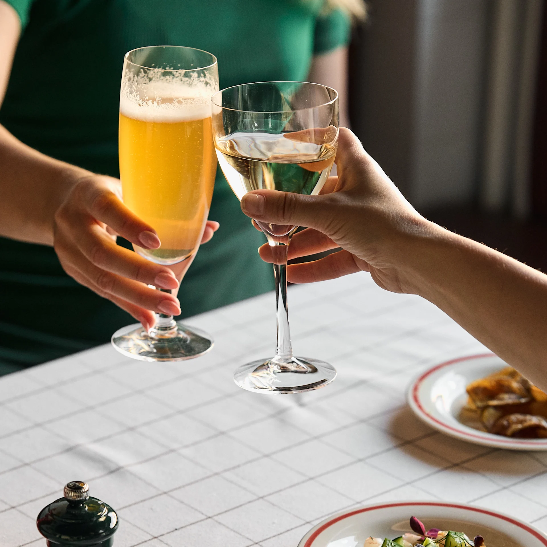 Two people toast with a glass of beer and a glass of white wine at a table with a white checkered tablecloth.
