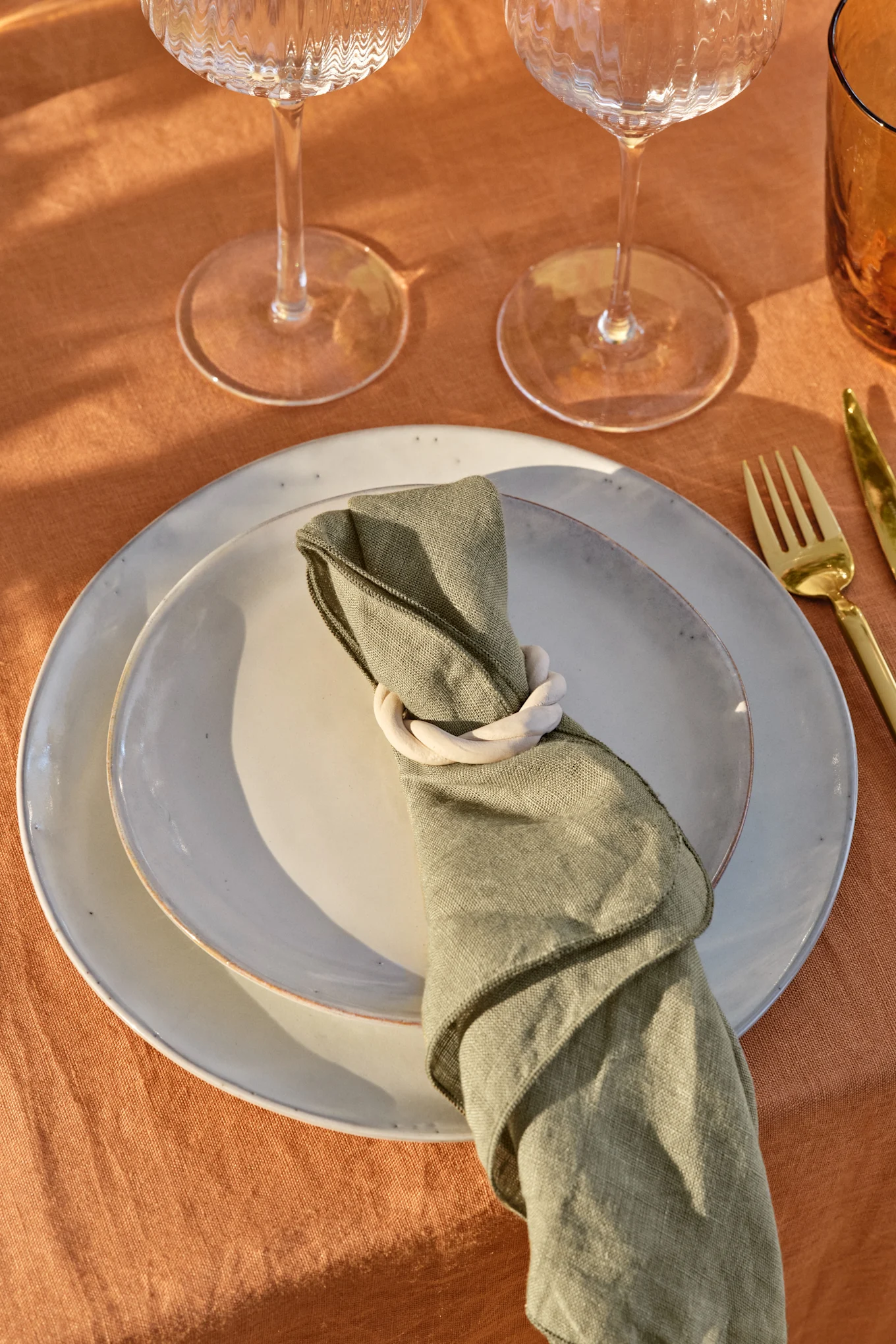 Table setting with light gray plates, olive green napkin with a white ring, gold cutlery, and glasses on a rust-colored tablecloth.