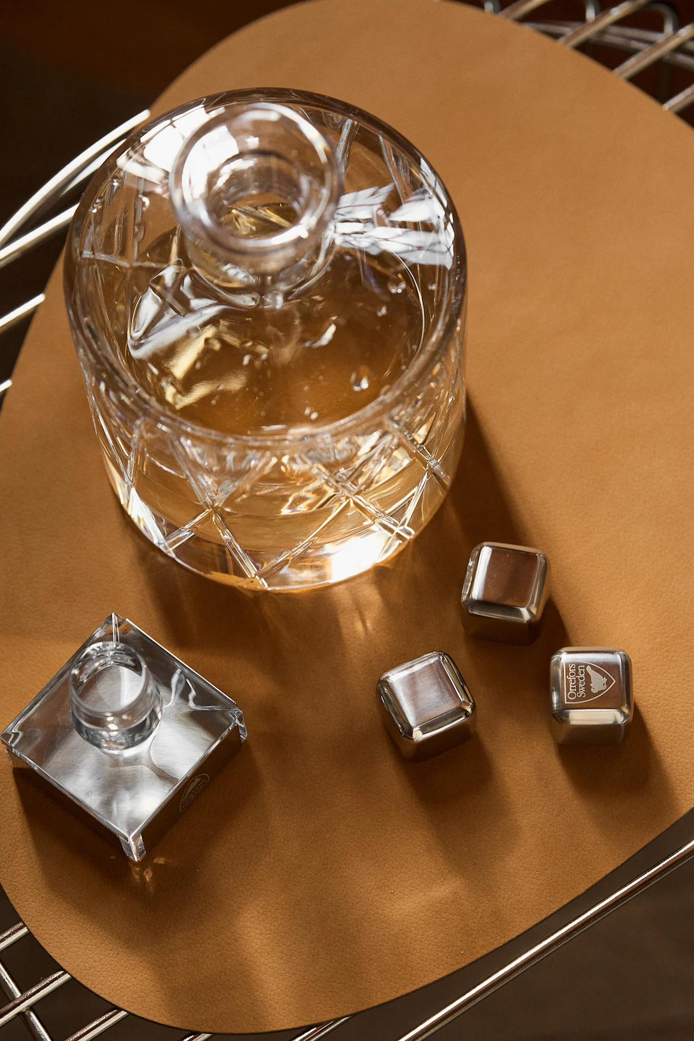 Crystal decanter with amber liquid, square crystal stopper, and three stainless steel chilling cubes on a round tan leather mat.