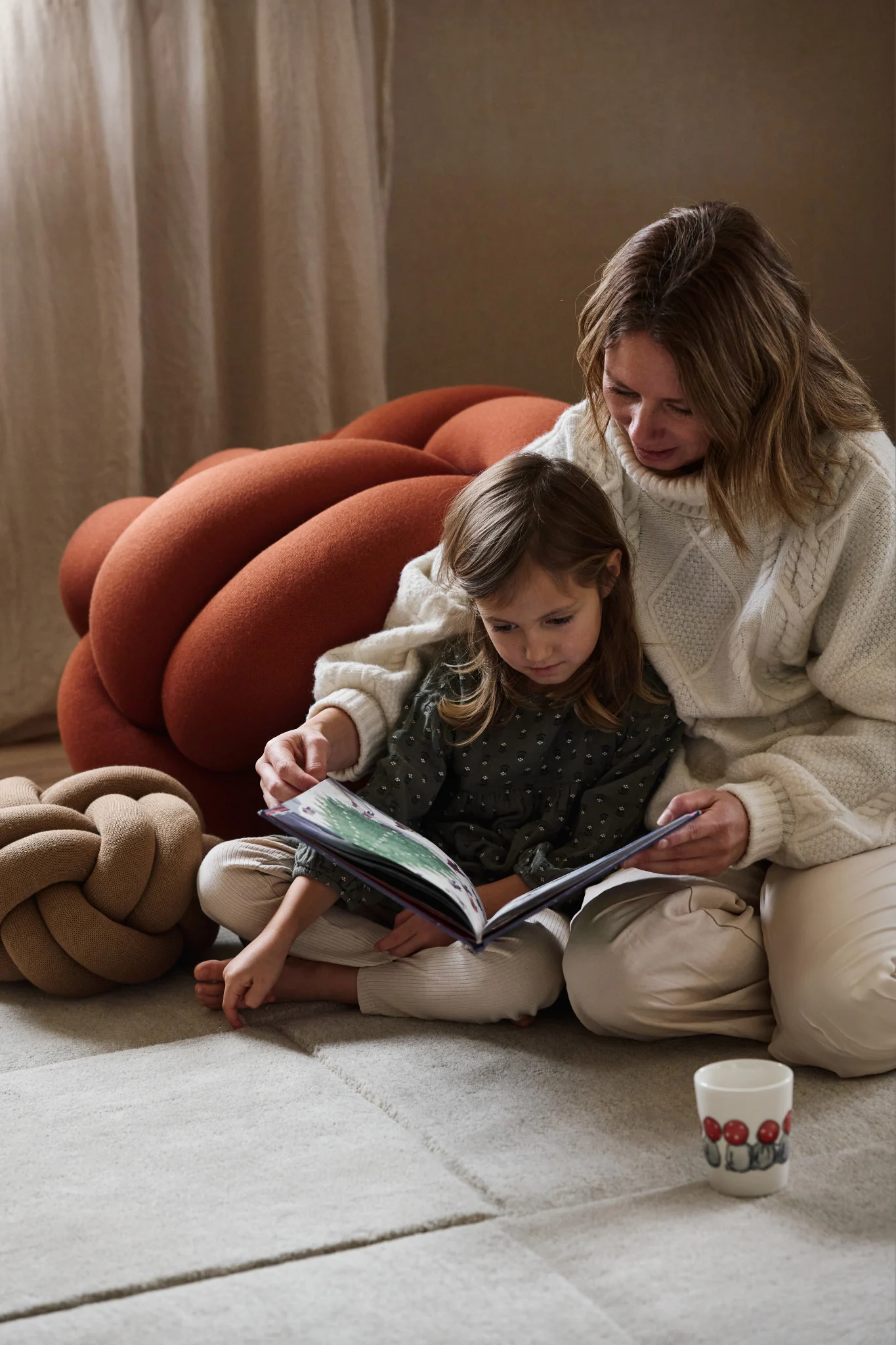 A woman and a girl sit on a rug, reading a book. A large rust-colored knot cushion is behind them, a smaller one to the left.
